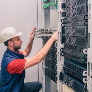 A man installs new equipment in a modern data center. An engineer in a white helmet mounts a stack of switches in a rack. The technician works in the server room. A man installs new equipment in a modern data center. An engineer in a white helmet mounts a stack of switches in a rack. The technician works in the server room.